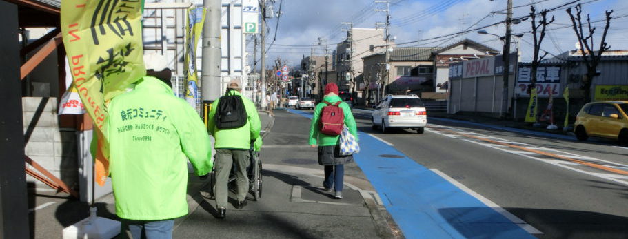 薄緑の揃いのジャンパーを着た3人が歩道を歩いている。一人は防災訓練の旗を持ち、一人は車椅子を押している。