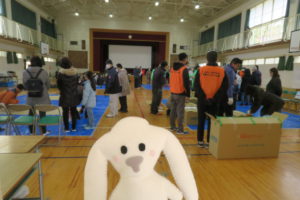 防災訓練の会場である安倉小学校の体育館の中に入ったまちキョン。その後ろにはすでに何人かの人が集まって、始まるのを待っている。