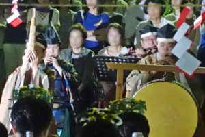 「祝詞（のりと）」に合わせて笛を吹く神事装束の人たち、その後ろには見学にきた人たちが写っています。手前右には太鼓が写っています。